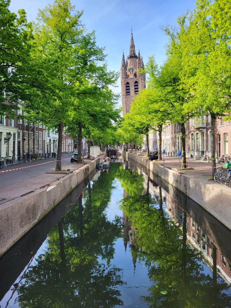 Old Church of Delft reflected in the water of a pristine canal lined with green trees