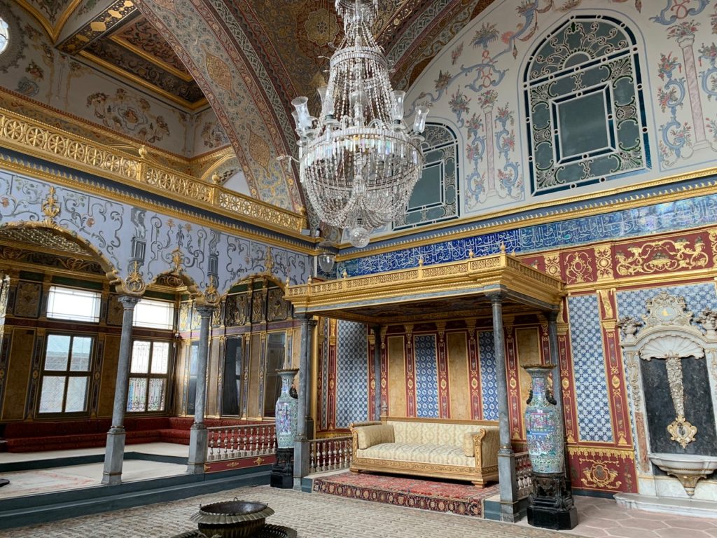 Crystal chandelier hangs in the middle of a blue, red, and gold chamber tiled with Arabic calligraphy and hosting a covered sofa