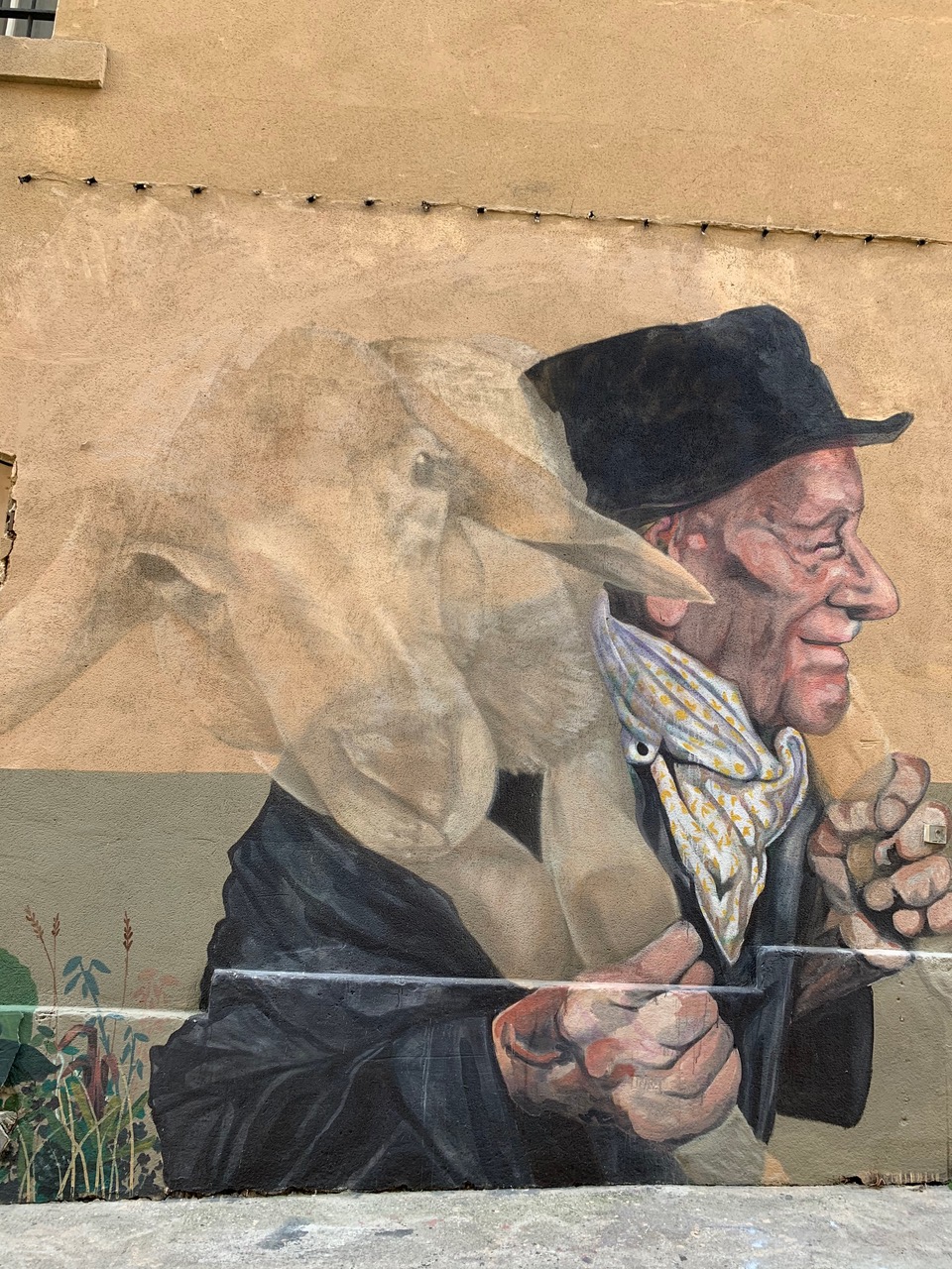 a painted elderly man with a black hat and jacket carrying a sheep over his shoulders