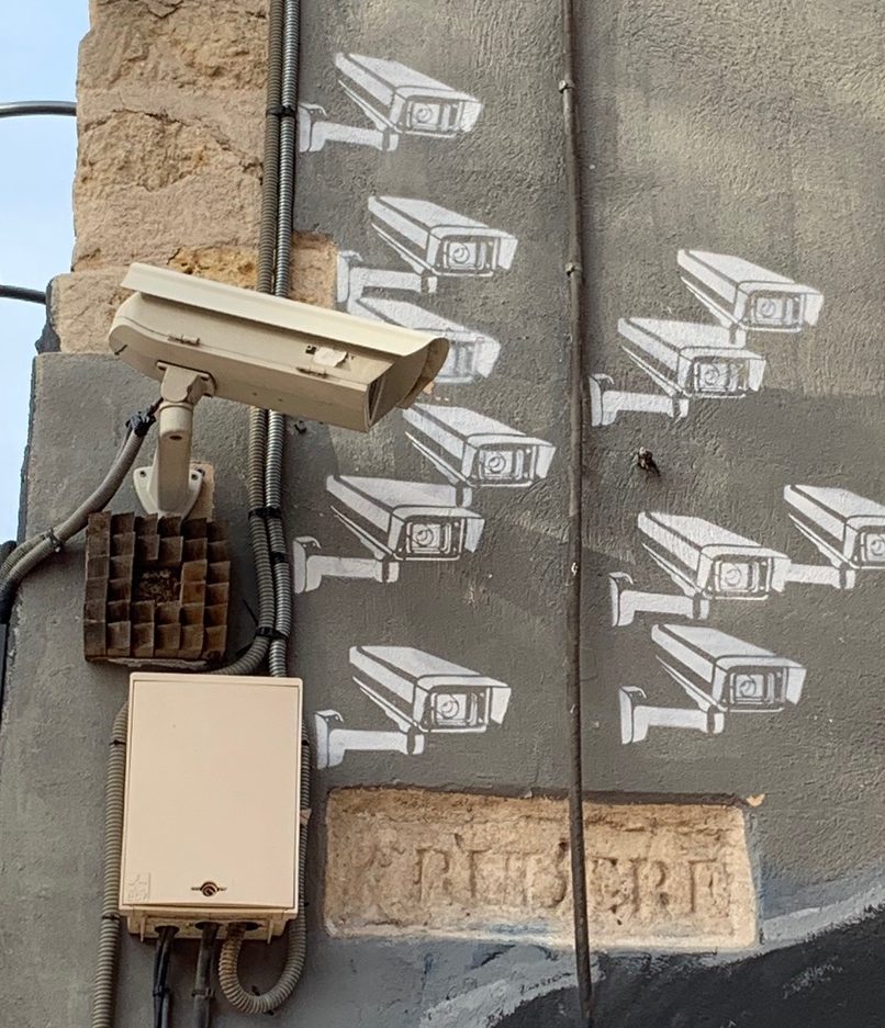 A security camera on a street corner with ten more cameras painted on the wall behind