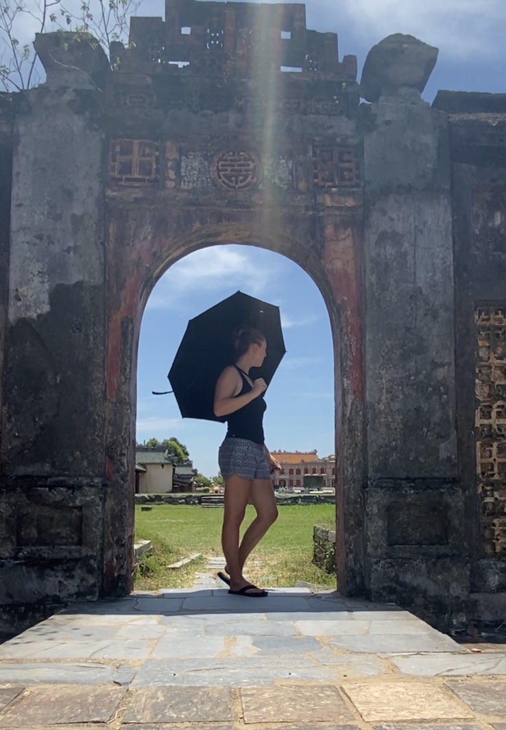 Maddie standing sideways with an umbrella over her shoulder framed by the curved archway of a wall in the old imperial palace of Hue